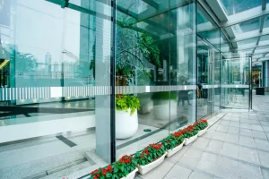 Modern glass building entrance with greenery and flowers.